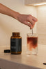 Person stirring a drink in a glass with a bottle labeled 'BIO BLANDS' on a countertop.