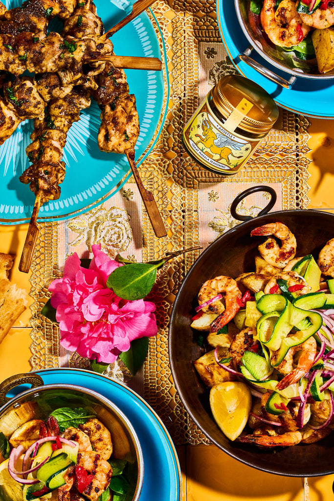 Assorted dishes including skewers, a bowl of salad, and a jar on a decorative tablecloth.