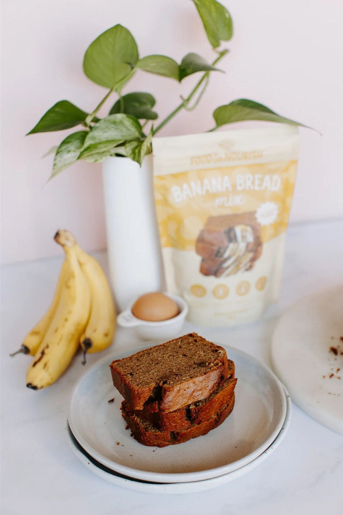 Two slices of banana bread on a plate with a package of banana bread mix, bananas, and an egg in the background.