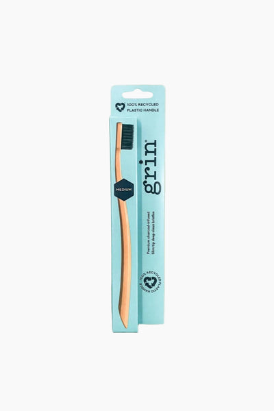 A medium toothbrush with orange bristles and a brown handle, packaged in a cardboard casing with 'grin' branding and product information.