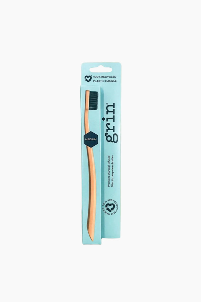 A medium toothbrush with orange bristles and a brown handle, packaged in a cardboard casing with 'grin' branding and product information.