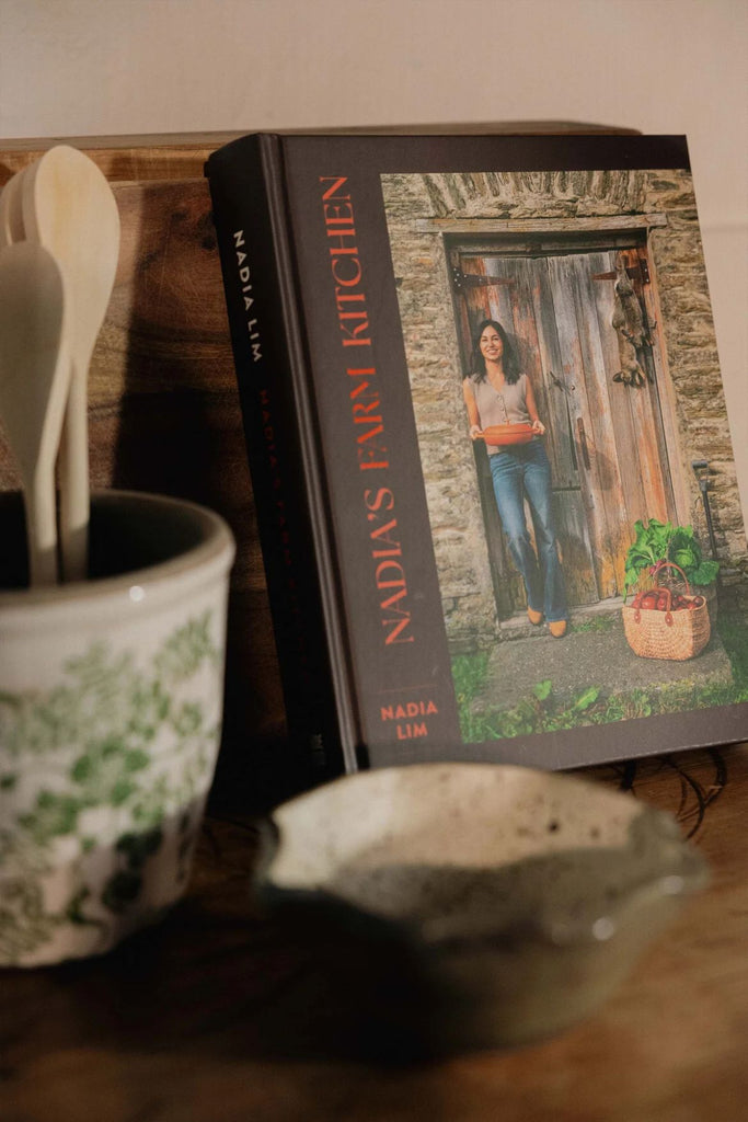 Cookbook titled 'Nadia's Farm Kitchen' on a wooden surface with a pot and spoon.
